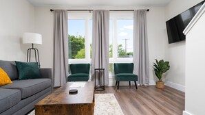 Un salon moderne avec un canapé gris, deux fauteuils verts, une table basse en bois et une grande fenêtre avec des rideaux.