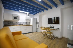 Cocina compacta con armarios blancos, un microondas y un refrigerador. Hay un sofá amarillo y una mesa de comedor con sillas en la zona de estar.