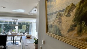 Et moderne spiseområde med et stort maleri på væggen, et bord med stole og udsigt til det fri gennem store vinduer.
