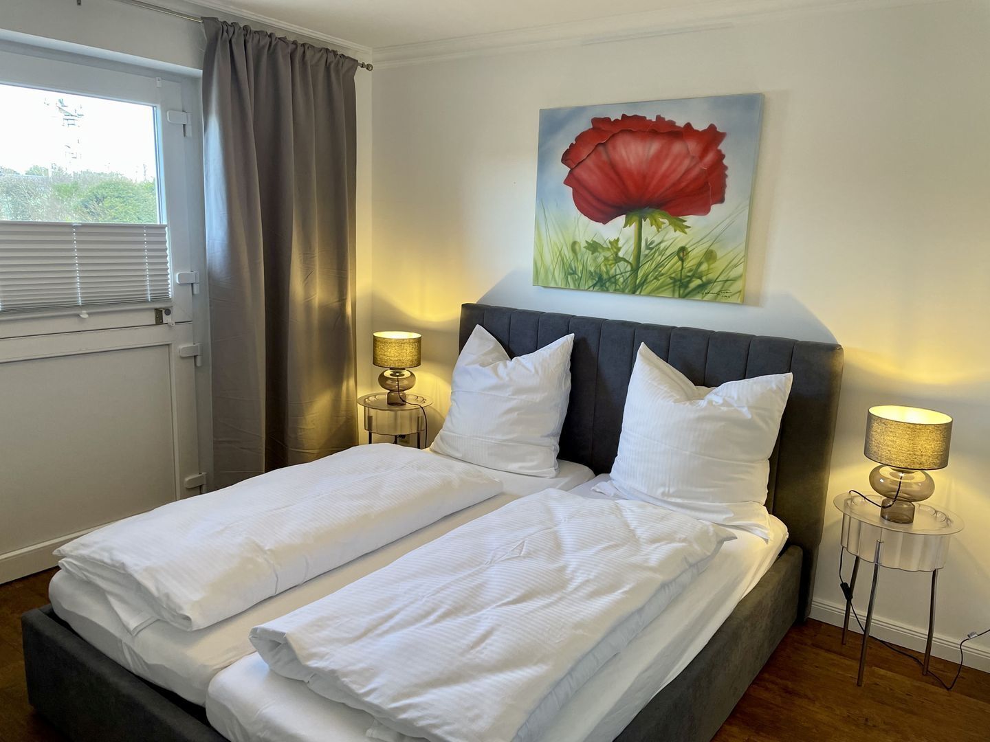 A double bed with white linens, two bedside lamps, a painting of a red flower on the wall, and a window with curtains.