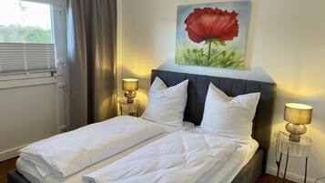 A double bed with white linens, two bedside lamps, a painting of a red flower on the wall, and a window with curtains.
