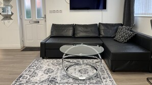 Uma sala de estar moderna com um sofá de couro preto, uma mesa de centro de vidro, um tapete estampado, uma TV de tela plana na parede e uma porta com painéis de vidro fosco.