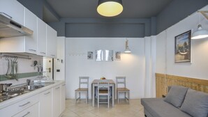 A compact kitchen with white cabinets, a dining table with chairs, a grey sofa, and a pendant light.