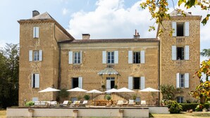 A large stone building with multiple windows and a central staircase. There are outdoor seating areas with umbrellas and lounge chairs. - Refurbished Chateau, Sleeps 20 with Luxury Pool (Labarthe)