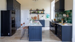 A modern kitchen with dark blue cabinets, a central island with stools, a built-in oven, and a variety of kitchenware on shelves. - Refurbished Chateau, Sleeps 20 with Luxury Pool (Labarthe)
