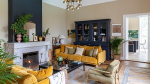 A living room with a fireplace, yellow sofa, dark blue cabinet, and a dining area visible through an open door. - Refurbished Chateau, Sleeps 20 with Luxury Pool (Labarthe)