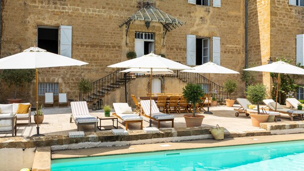 A poolside area with lounge chairs and umbrellas, a stone building with windows, and a staircase leading to an upper level. - Refurbished Chateau, Sleeps 20 with Luxury Pool (Labarthe)