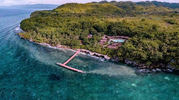 Vue aérienne d’une île tropicale avec une plage de sable fin, un quai et un complexe hôtelier aux bâtiments surmontés de toits de chaume.