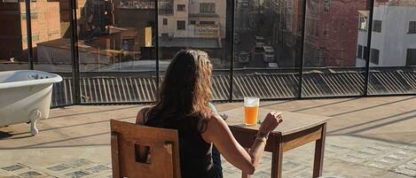 Una persona sentada a una mesa en una terraza en la azotea con vistas a la ciudad, una bañera y un cubo de basura.