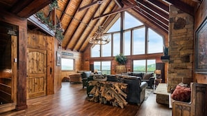 A spacious living room with a high ceiling, wooden beams, and large windows offering a view of the outdoors.
