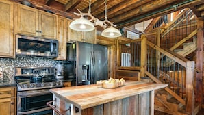 A rustic kitchen with wooden cabinets, a large island, stainless steel appliances, and a wooden staircase.