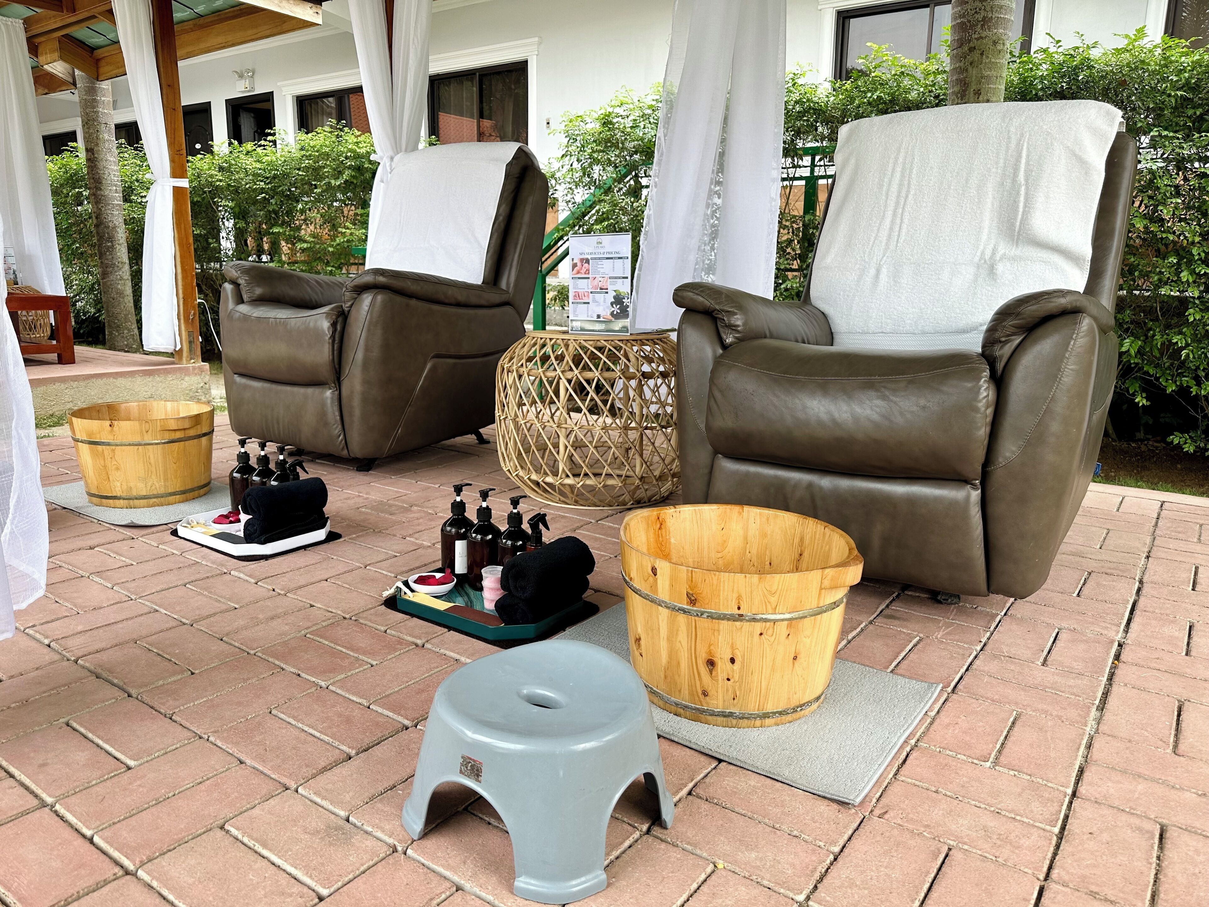 Two leather recliners with white covers, a wooden bucket, a small stool, and bottles of toiletries on a brick patio.