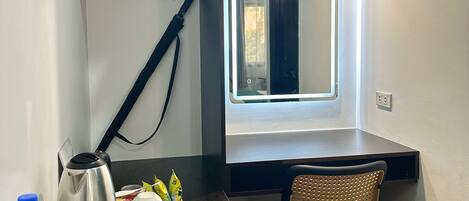 A compact hotel room with a small desk, a chair, a kettle, a water bottle, and a tray with two cookies.