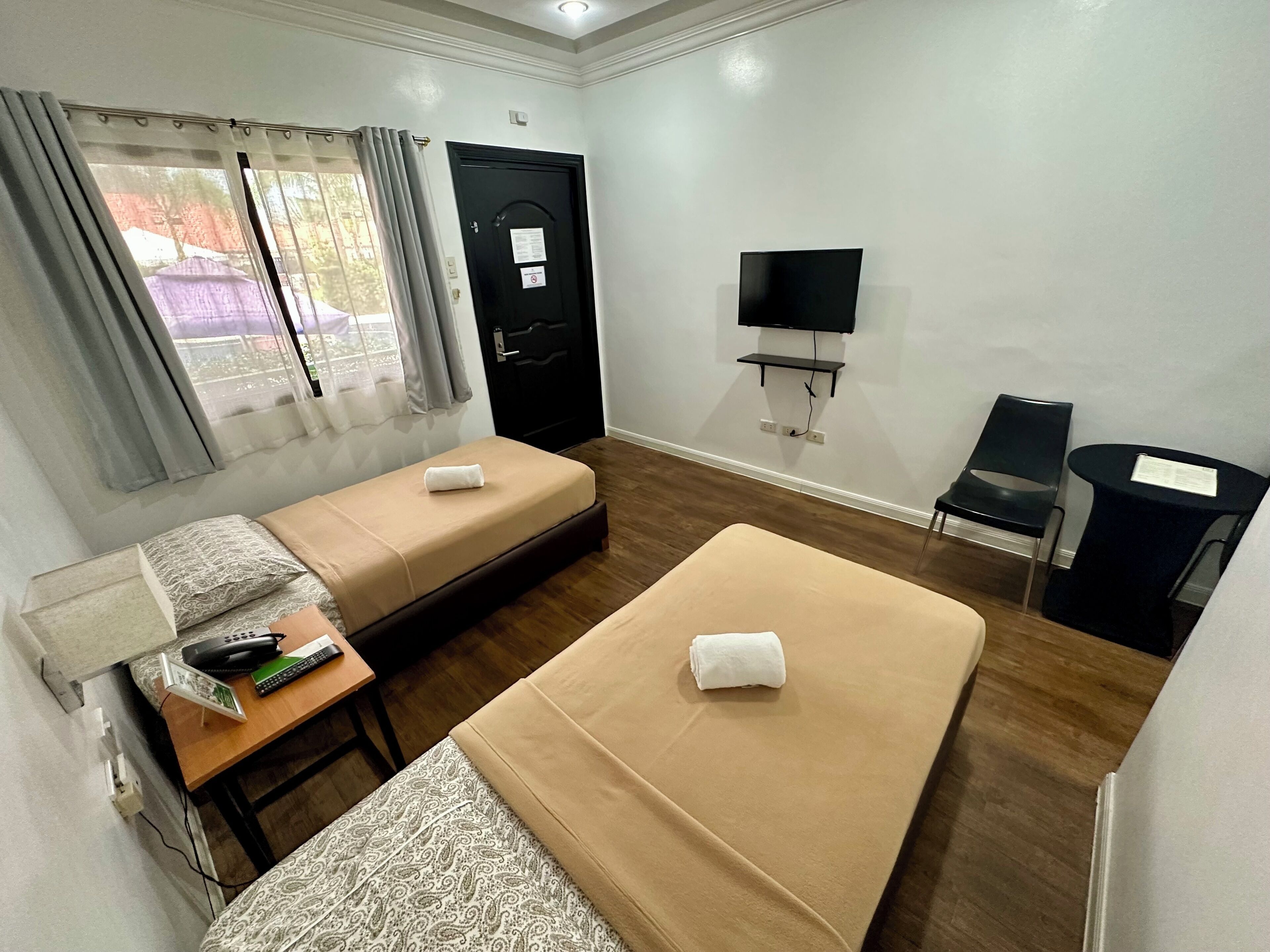 Ein Zimmer mit zwei Einzelbetten, einem kleinen Tisch mit Telefon und Fernbedienung, einem an der Wand befestigten Fernseher und einem Sessel.