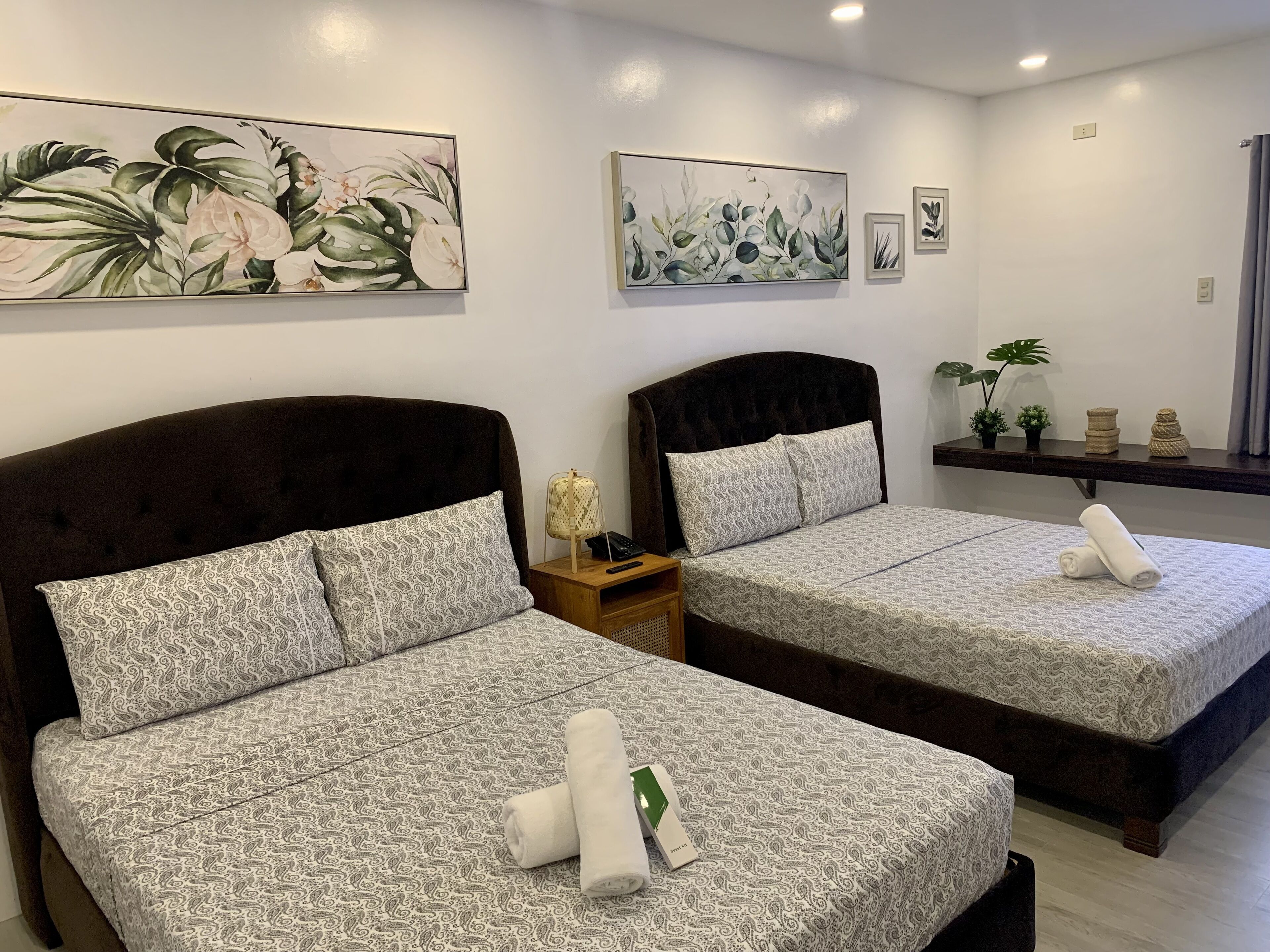 Dos camas con cabeceras a juego y ropa de cama estampada, una mesita de noche con lámpara y cuadros en la pared sobre las camas.