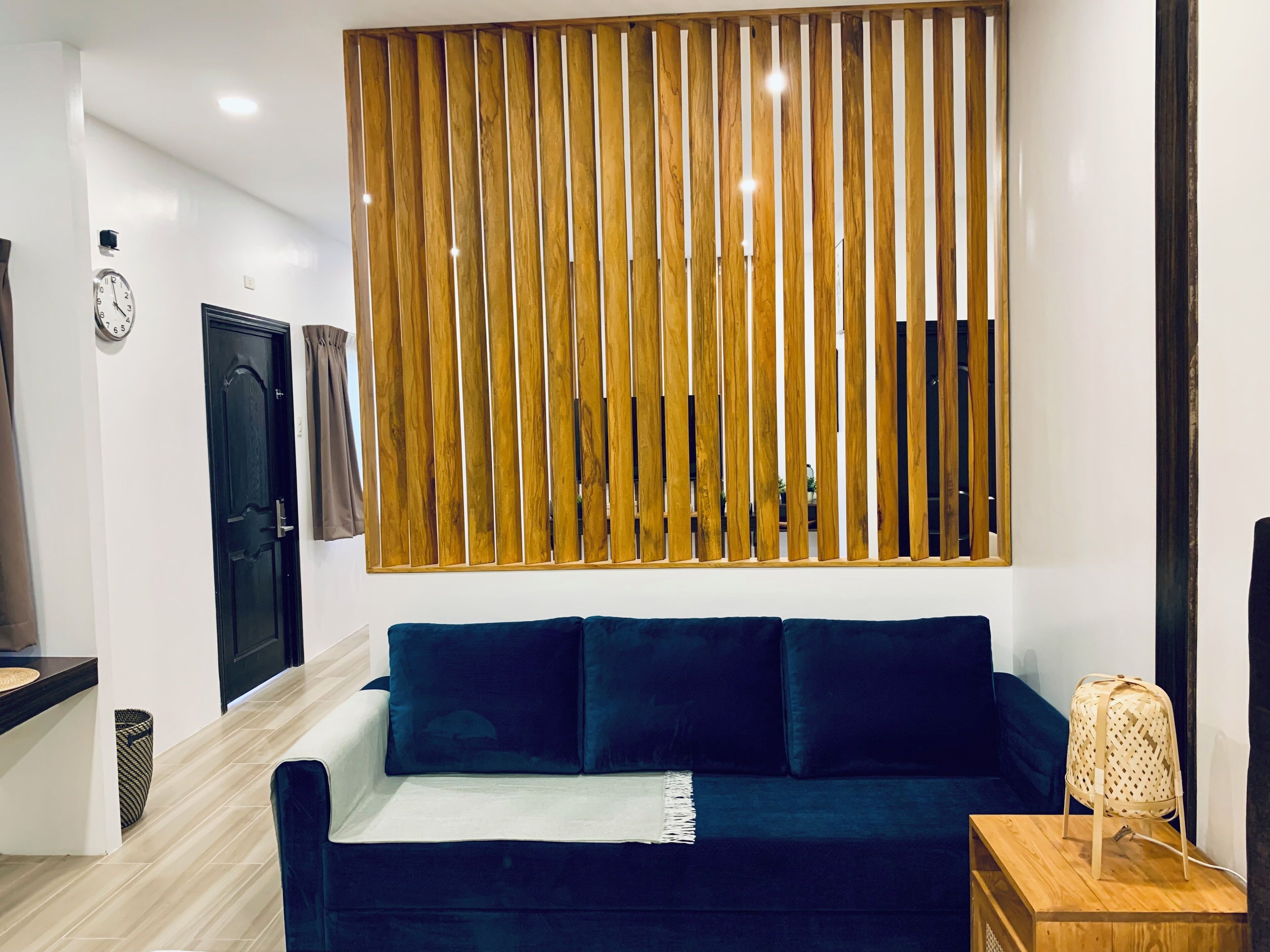Una sala moderna con un sofá azul oscuro, un panel de pared de listones de madera, un reloj y una pequeña mesa de madera con una cesta tejida.