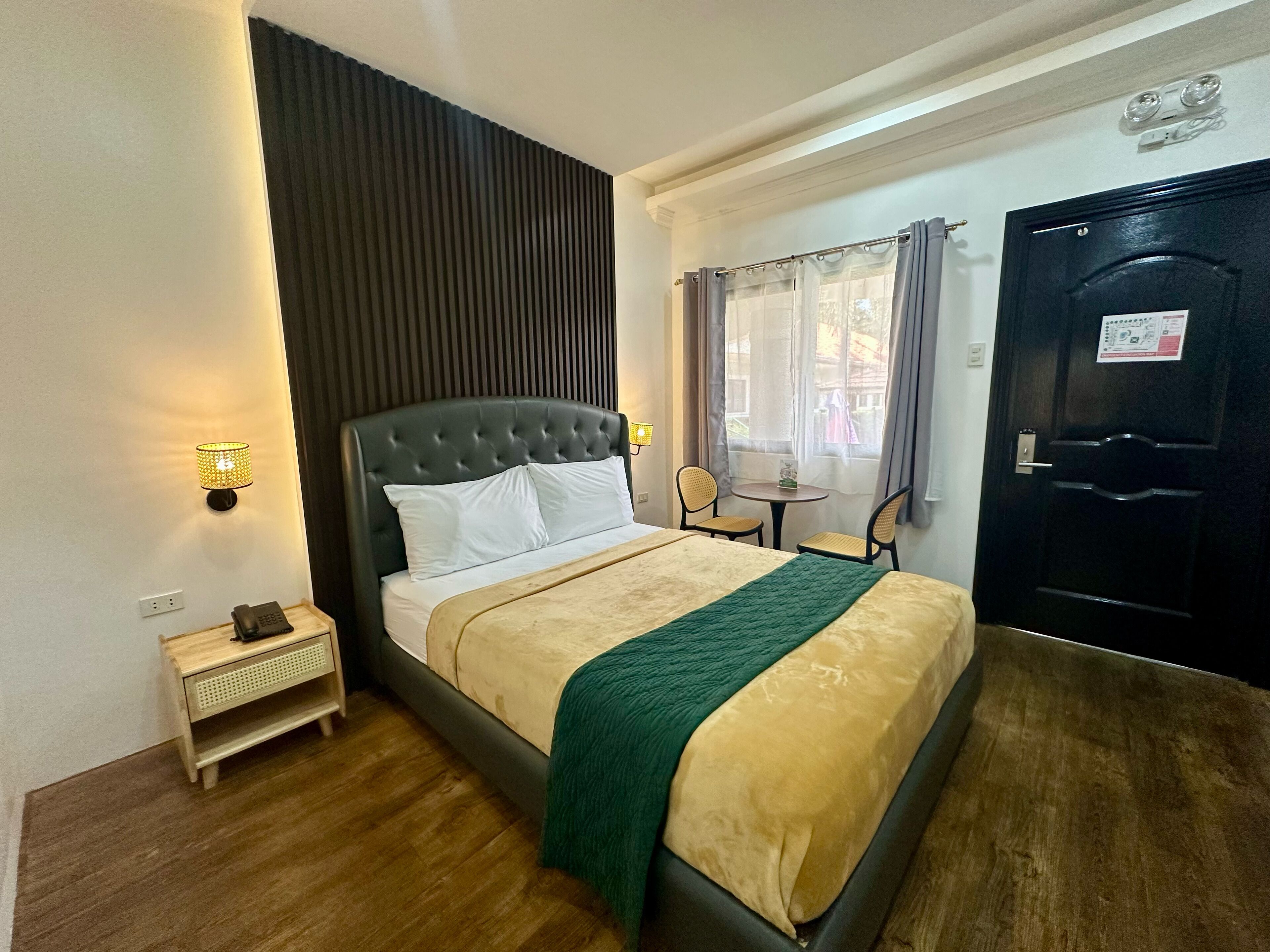A neatly arranged hotel room with a large bed, a bedside table with a lamp, a small table with chairs, and a door with a sign.