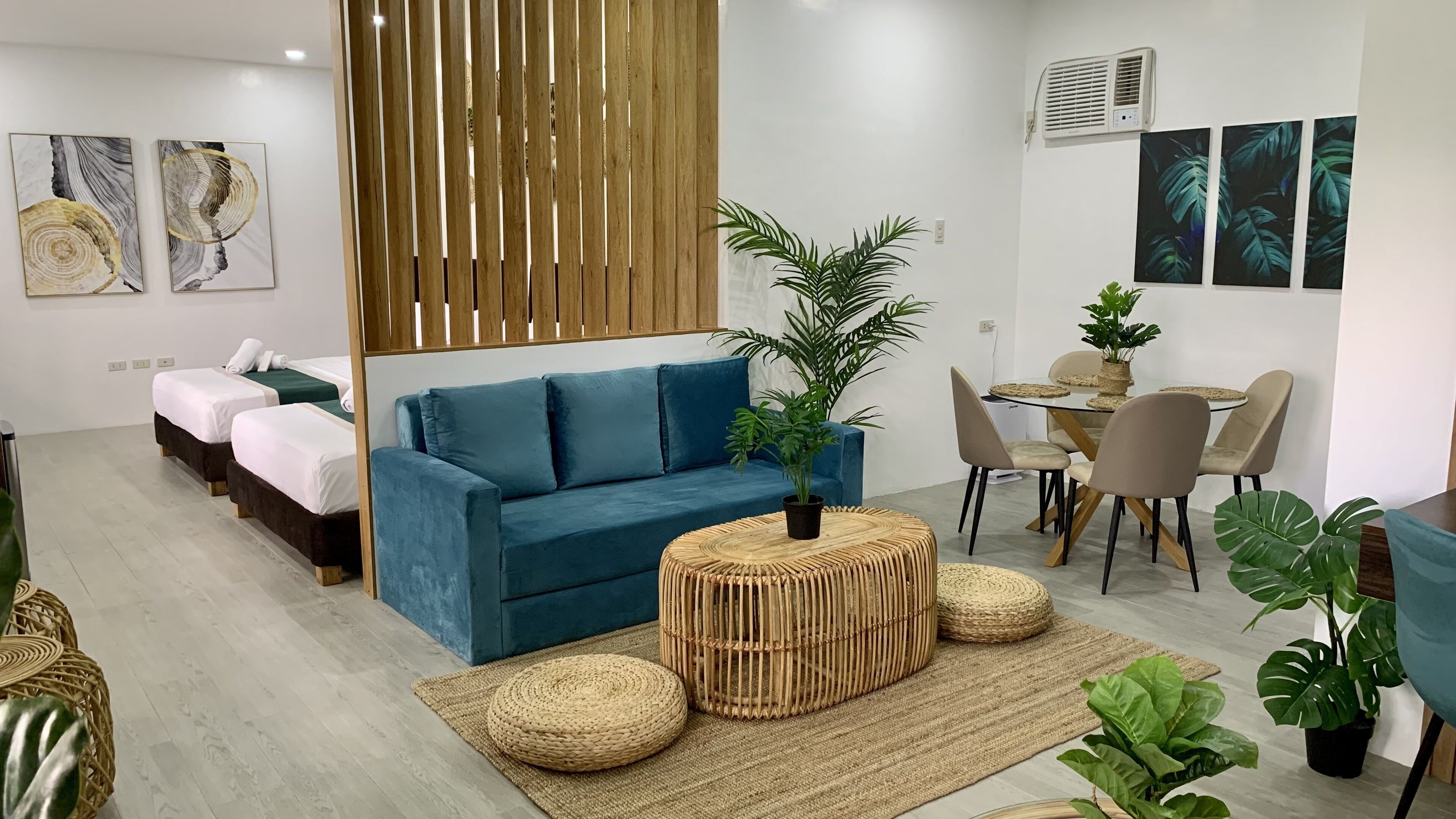 Una habitación de hotel moderna con un sofá azul, dos camas individuales, un comedor y varias plantas en macetas.