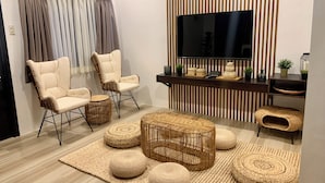 A modern living room with a flat-screen TV, two white cushioned chairs, a wooden coffee table, and woven poufs on a beige rug.