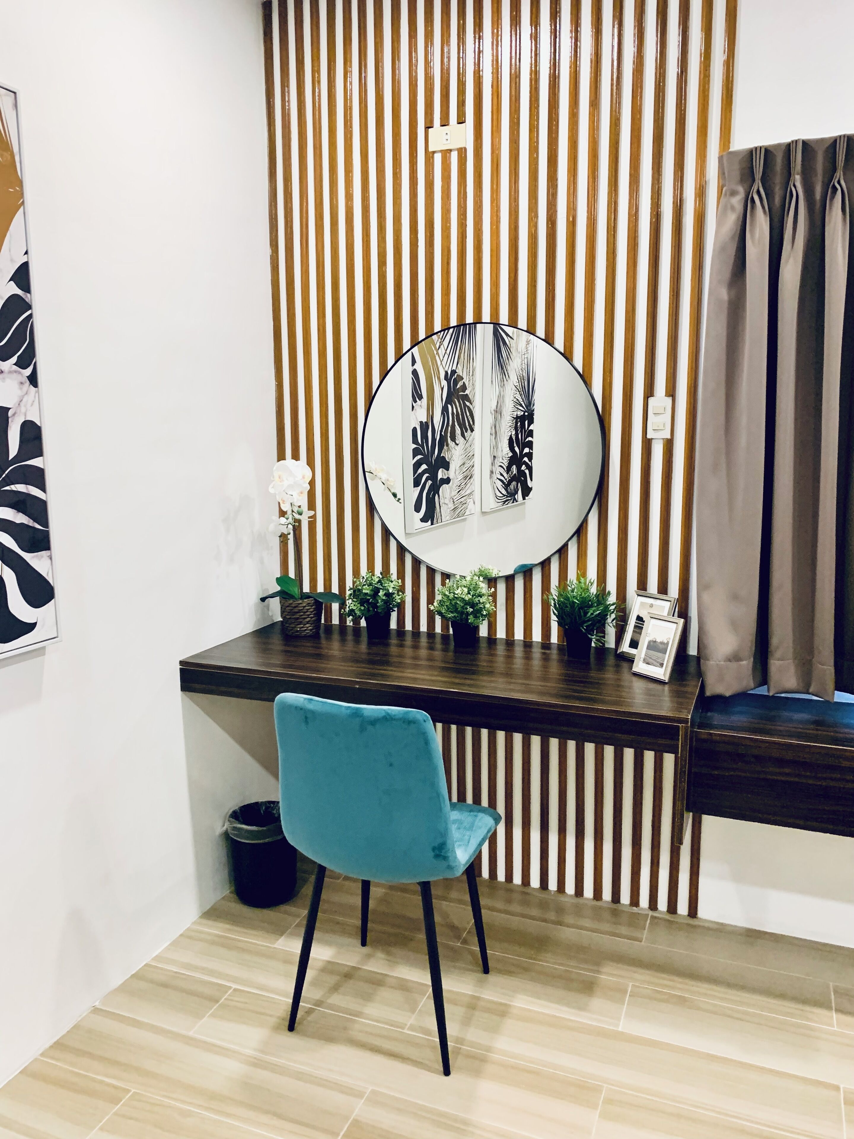 A corner room with a wooden desk, a blue chair, a round mirror, and a striped wall.