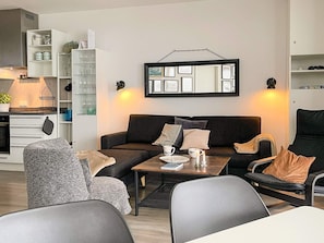 A modern living room with a sofa, armchairs, and a coffee table. There is a bookshelf with glass doors and a kitchen area in the background. - Sandpiper (Wendtorf)