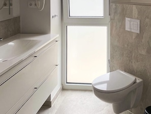 A modern bathroom with a white sink, vanity, and toilet. There is a window above the toilet, and a towel rack is visible on the wall.
