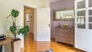 A hallway with a wooden floor, a white door, a mirror, a dresser with decorative items, and a large plant.