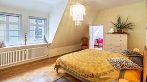 A bedroom with a sloped ceiling, a chandelier, a bed with patterned bedding, a dresser, a plant, and a window with a view.
