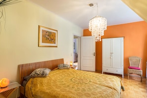 A bedroom with a bed, a chandelier, a wardrobe, a chair, and a picture on the wall.