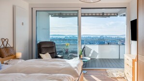 Une chambre avec un lit, une chaise, une petite table avec un vase, un balcon avec vue et un téléviseur fixé au mur.