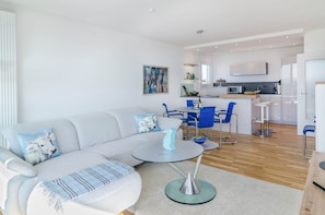 Ein modernes Wohnzimmer mit einer Couch, einem Couchtisch und blauen Sesseln. Im Hintergrund ist eine Küche mit Mikrowelle und Spüle erkennbar.