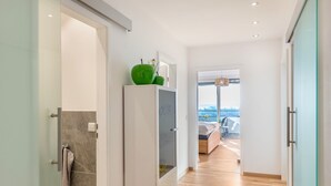 Un couloir avec un plancher en bois, une armoire blanche sur laquelle repose un objet vert, et un lit avec une vue en arrière-plan.