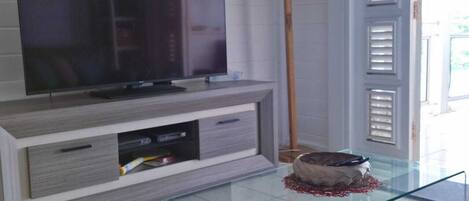 Una sala moderna con un televisor de pantalla plana sobre un mueble de madera, una mesa de centro de vidrio, una lámpara de pie y vistas al exterior a través de una ventana con persianas.
