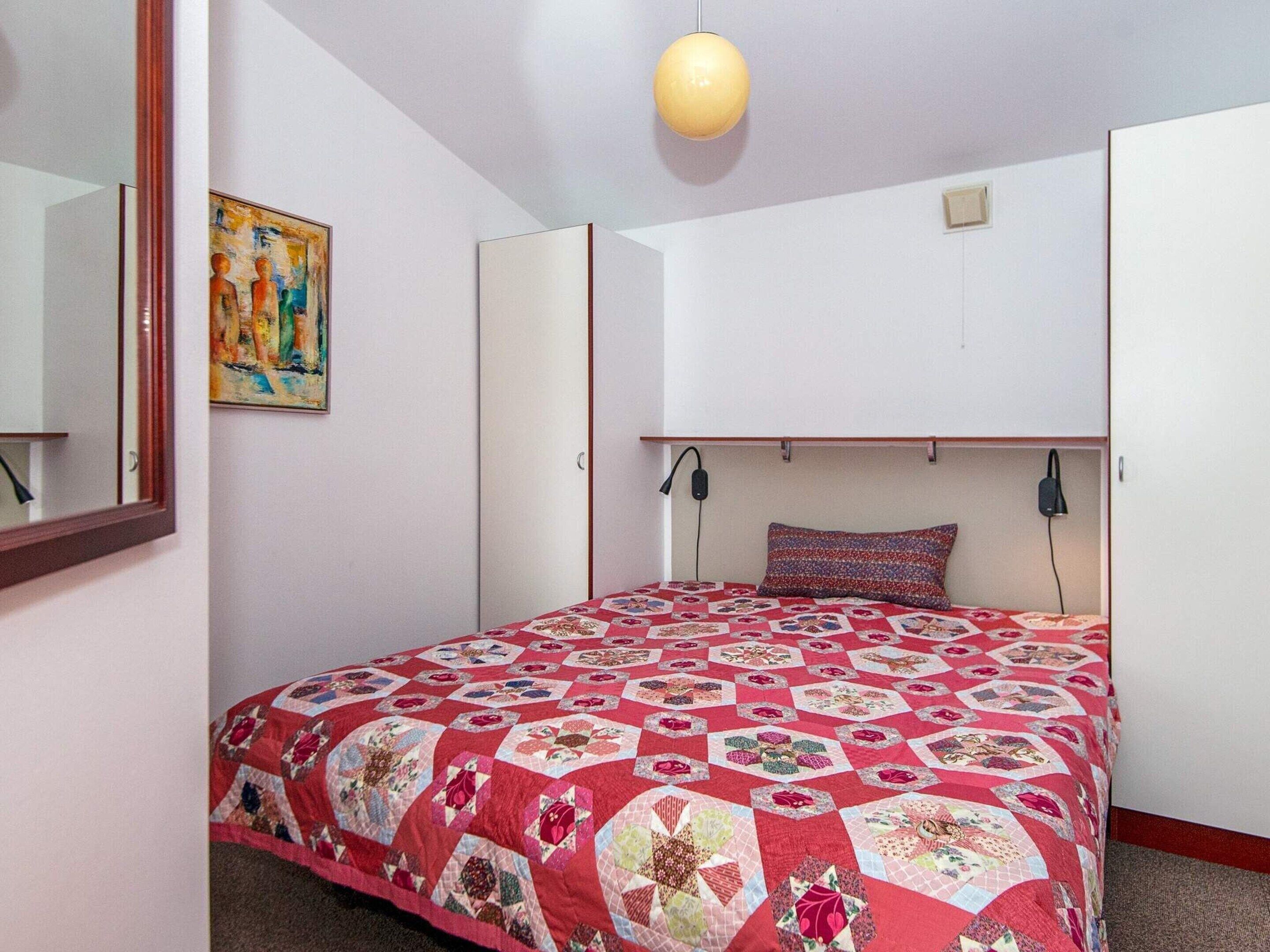 A bedroom with a red quilted bedspread, a painting on the wall, and a ceiling light.