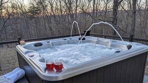 Een hottub met twee glazen rode drank en een kaars, omringd door een houten terras en een bebost achtergrond.