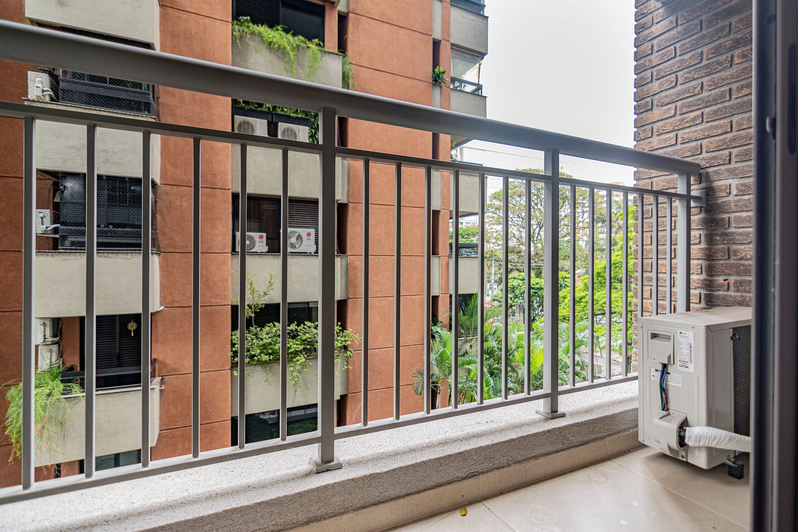 A balcony with a metal railing overlooking a brick building with multiple air conditioning units and potted plants.