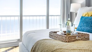 A hotel room with a large window, a bed with white bedding, a woven tray with a bottle and glasses, and a view of the sea.