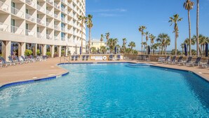 A hotel pool with sun loungers, palm trees, and a high-rise building in the background.