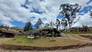 A rustic thatched-roof building labeled "ESTACIONAMENTO" surrounded by trees and a cobblestone path. - PONDOWASI LODGE (Angochagua)