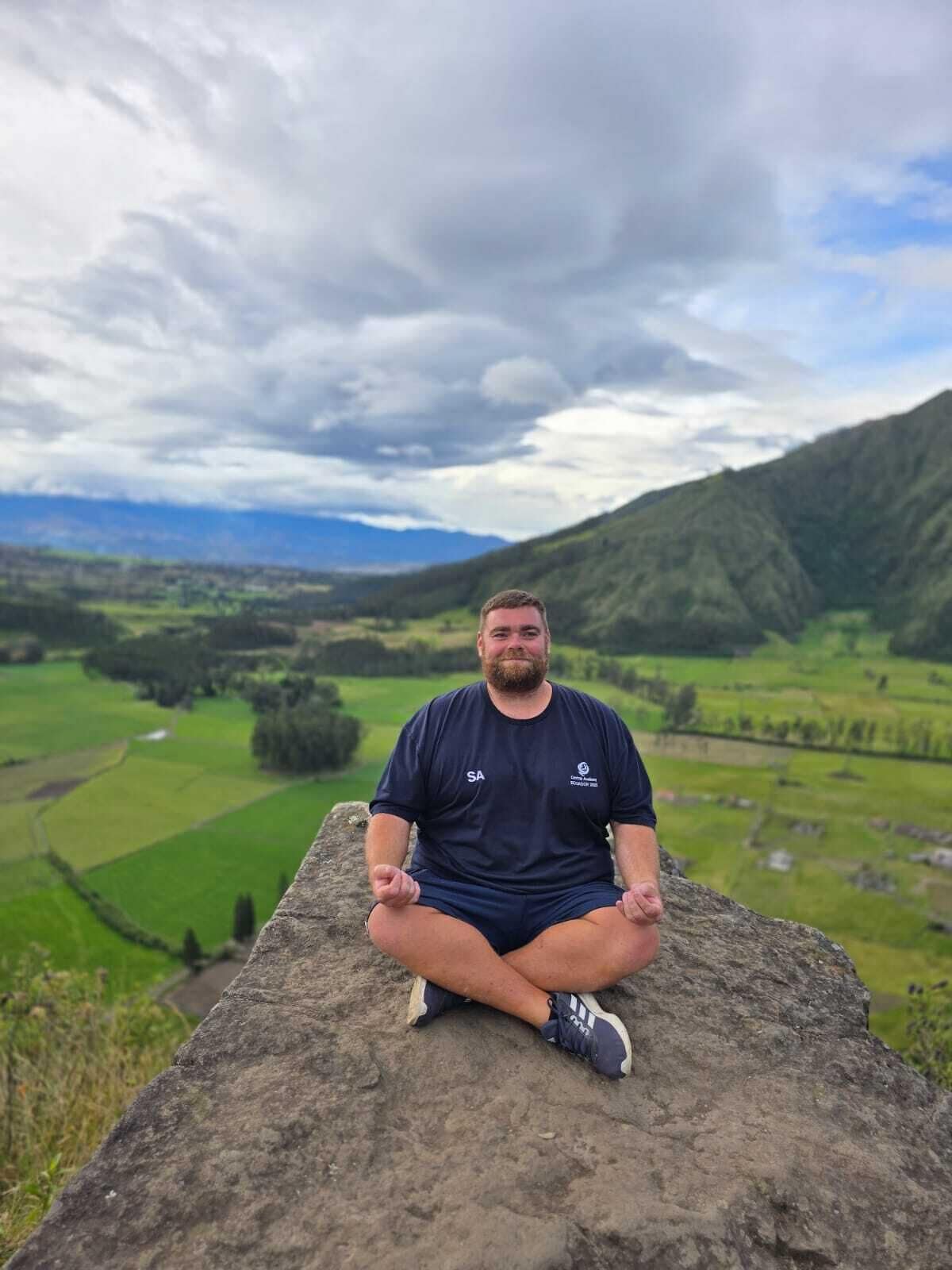Une personne assise sur un rocher, avec une vue panoramique sur des champs verdoyants et des montagnes en arrière-plan.