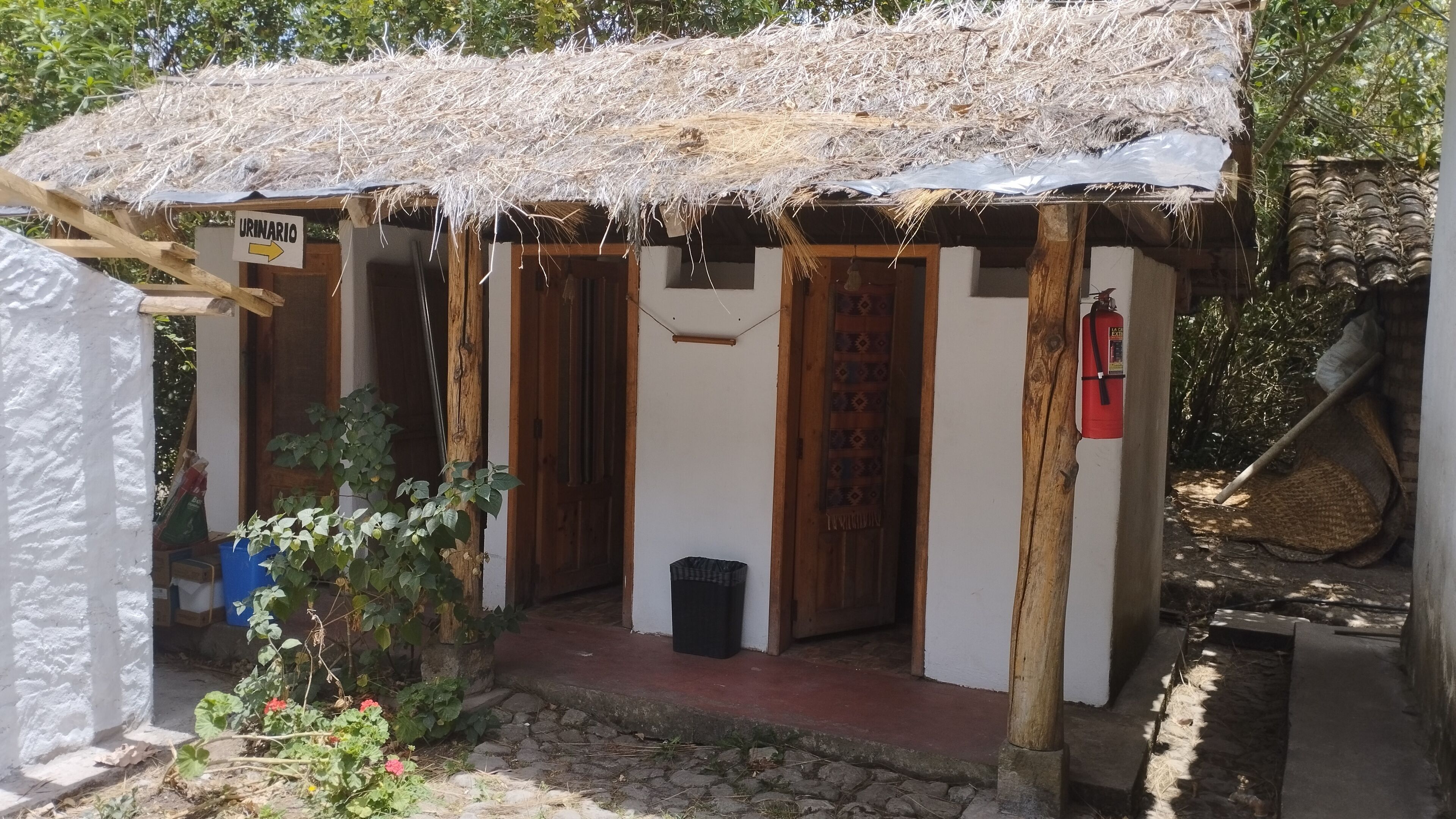 A small, rustic building with a straw roof, wooden doors, and a sign indicating a urinal.
