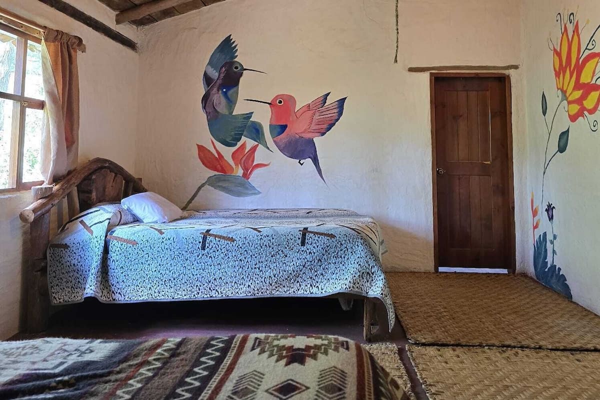 Quarto rústico com teto de madeira, cama com roupa de cama estampada, um mural de aves e uma porta.