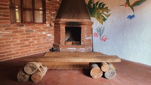 A rustic brick fireplace with a chimney, a wooden table with logs, and a mural of a hummingbird on the wall. - PONDOWASI LODGE (Angochagua)