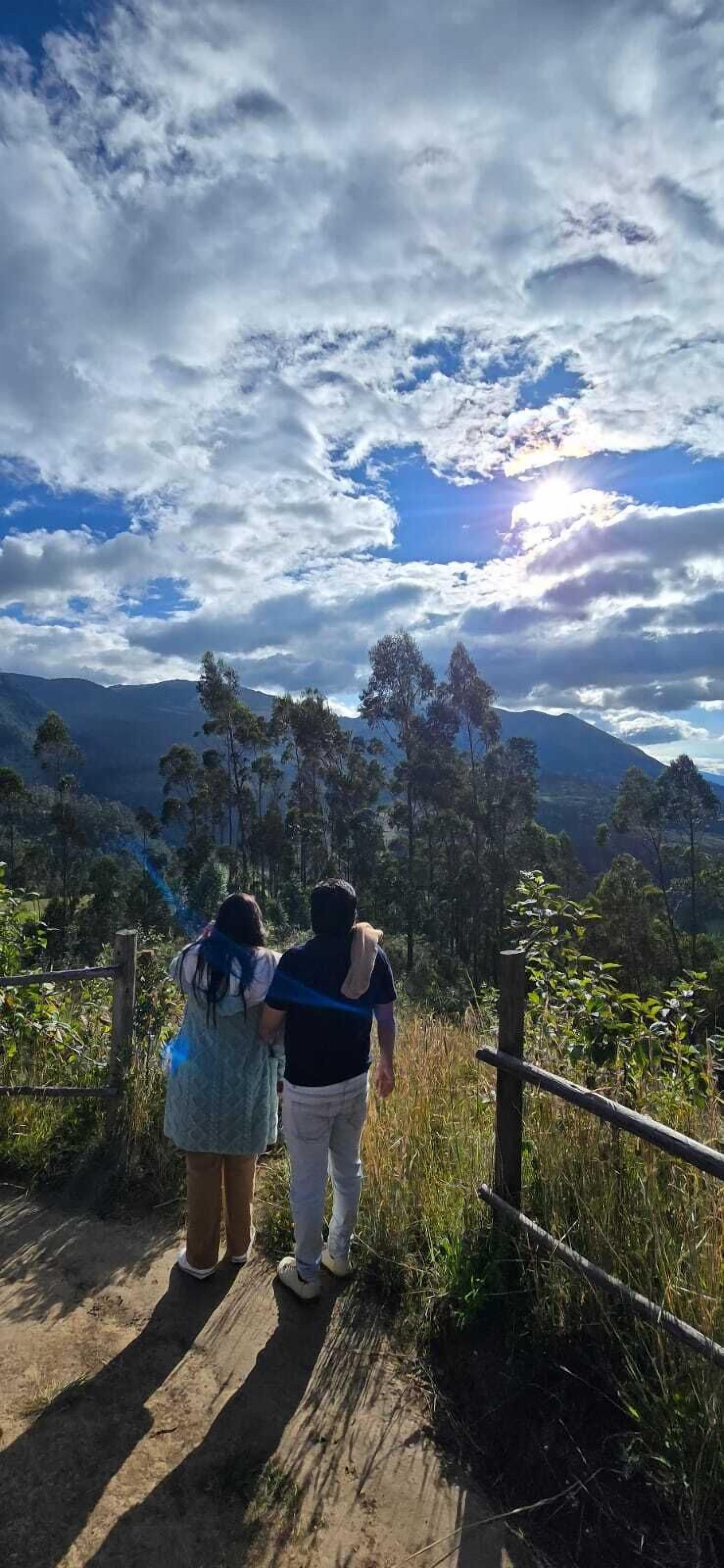 A couple standing on a wooden pathway with a scenic view of mountains and trees under a partly cloudy sky.