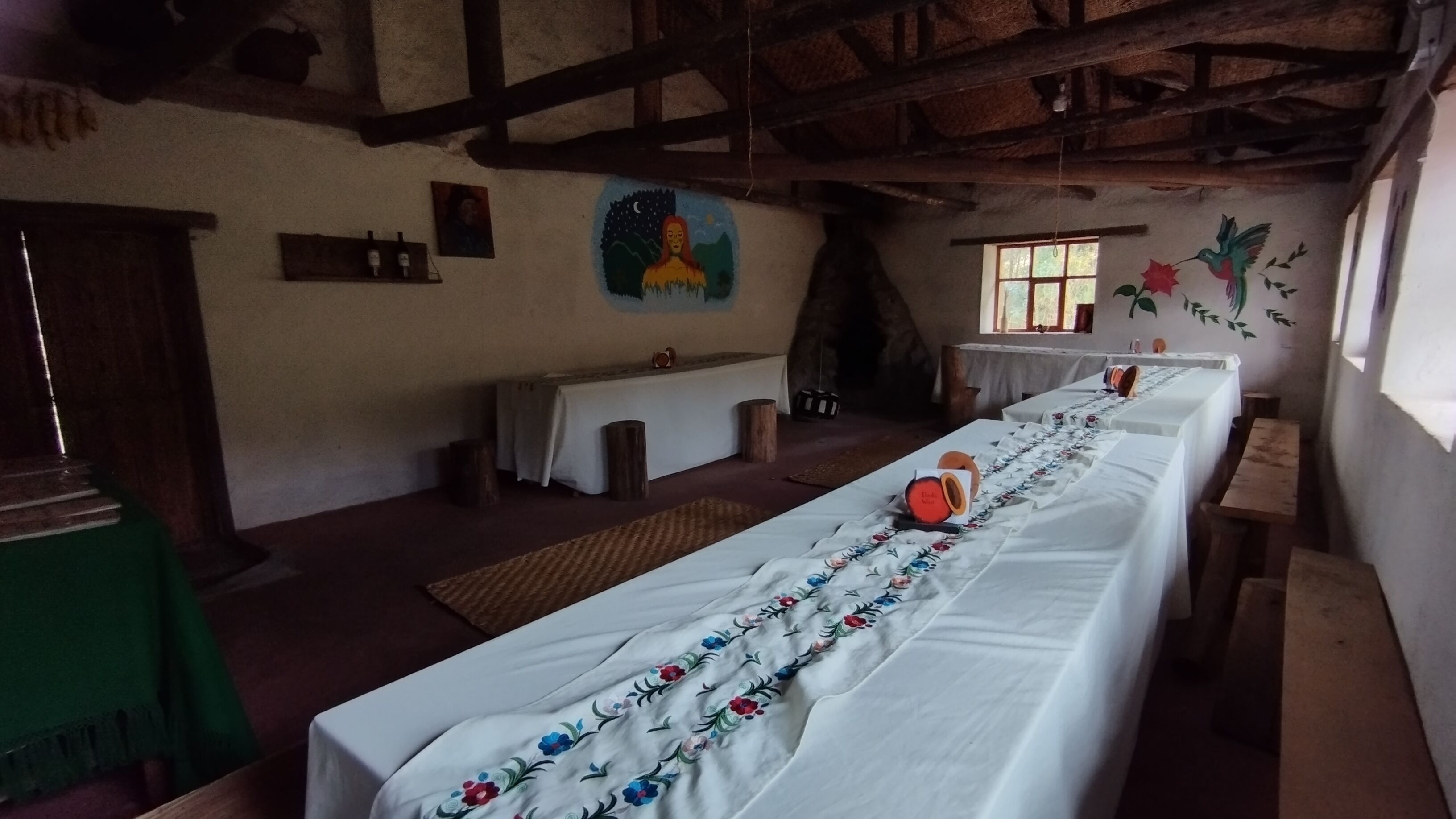 Une longue table à manger recouverte de nappes à motifs floraux, des bancs en bois et un intérieur rustique avec des poutres apparentes et une fresque murale.