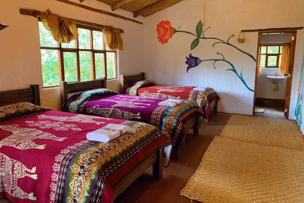 Quarto com duas camas, janela com cortinas, painel na parede e porta de banheiro.