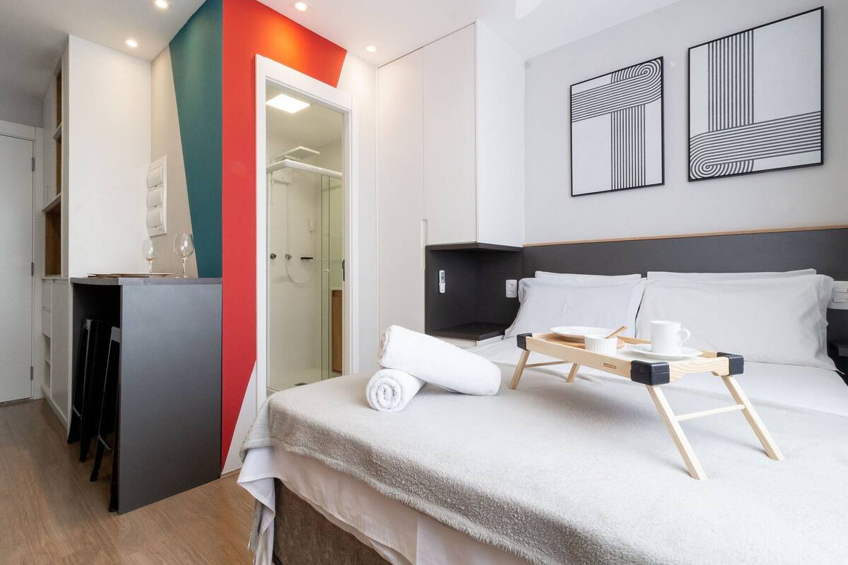 Hotellihuoneessa on sänky, yöpöytä ja pieni työpöytä tuolilla. Sängyllä on pyyhe ja pöydällä tarjotin, jossa on kuppi ja aluslautanen.