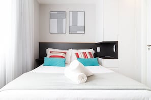 Cama bem arrumada com roupa de cama branca e duas almofadas, cabeceira preta e televisão fixada na parede.