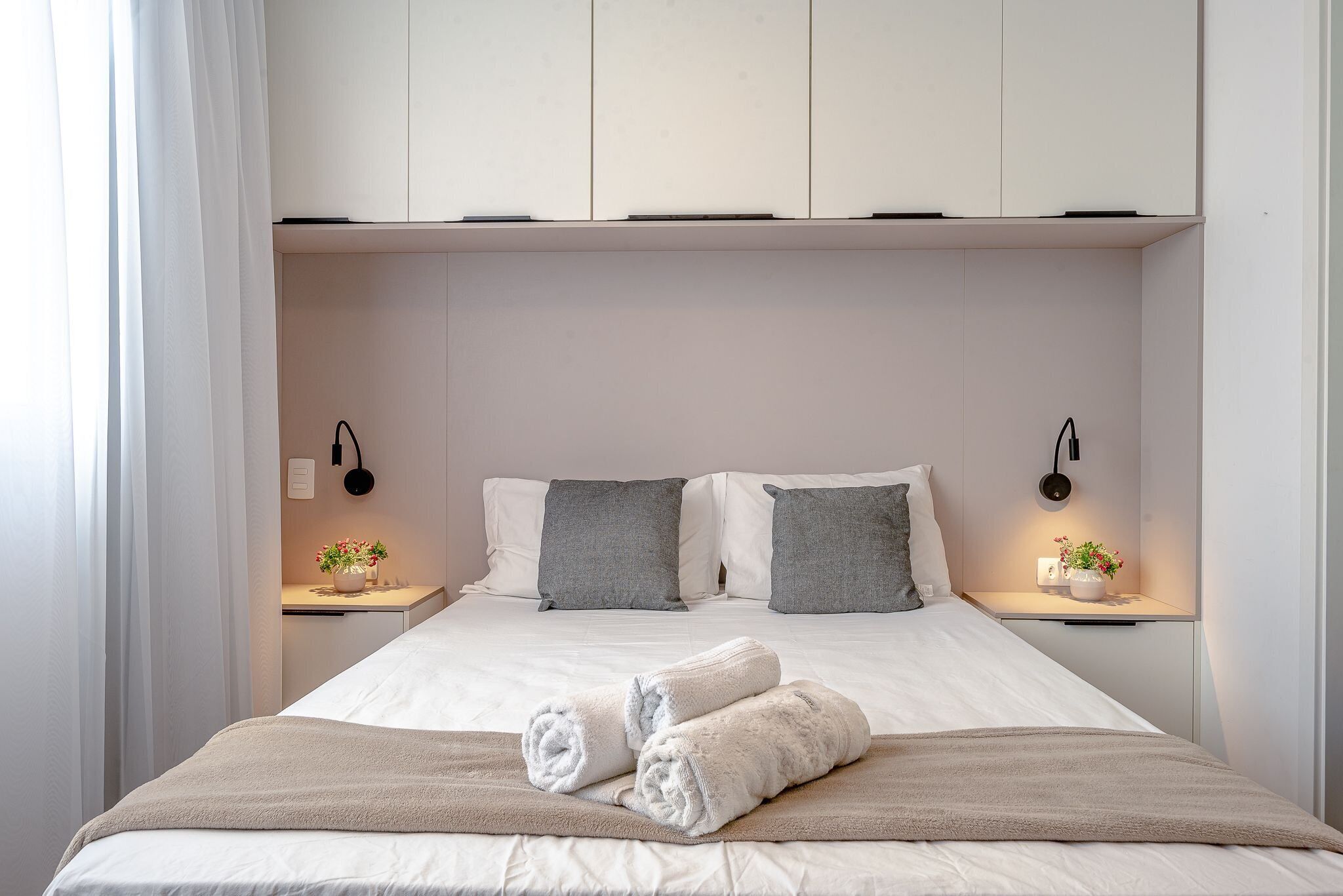 A neatly made bed with white linens and two rolled towels, a bedside table with a small plant, and a wall-mounted lamp.