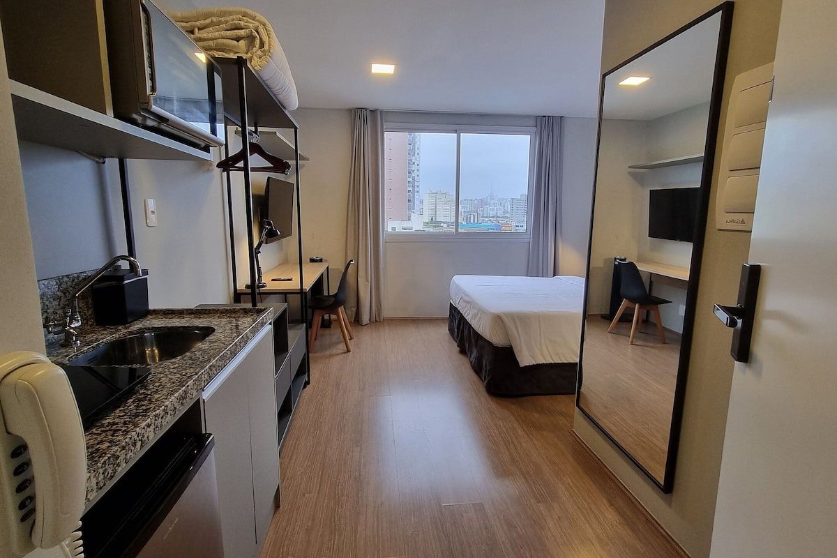 Kompakti hotellihuone, jossa on pieni keittiönurkkaus, sänky, työpöytä ja suuri peili.